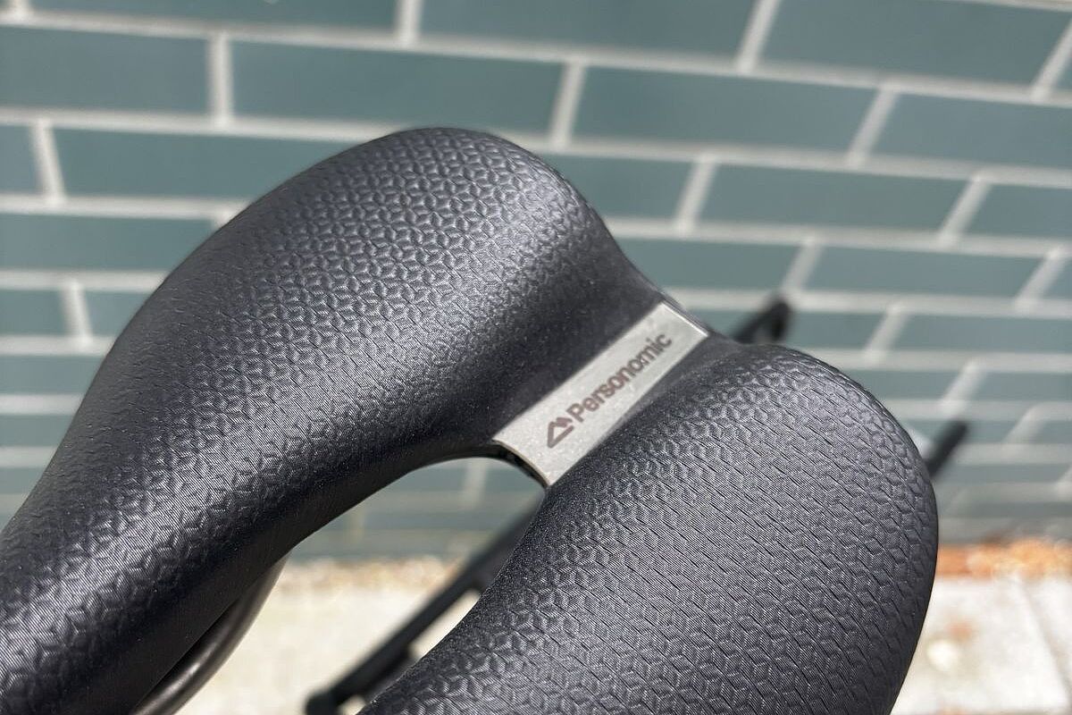 Detail eines schwarzen Fahrradsattels mit feiner Struktur und Metallplakette vor hellgrüner Kachelwand.